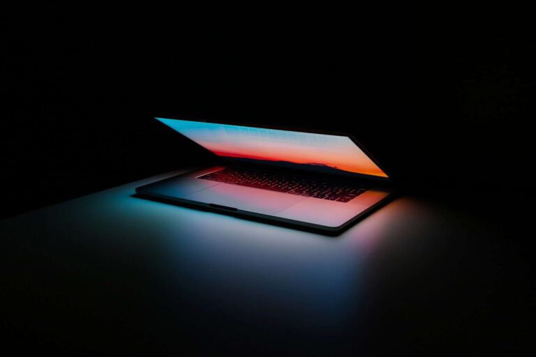 A partially open laptop glows in a dark room, casting colorful light across the surface. The screen displays a gradient of warm colors, creating a contrast against the surrounding darkness.