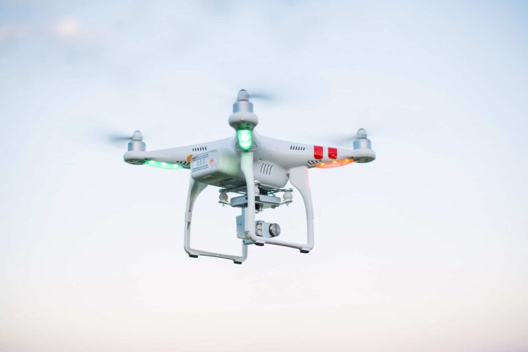 A white quadcopter drone with red and green lights hovers in a clear sky. Its camera is visible underneath, capturing aerial footage or images.