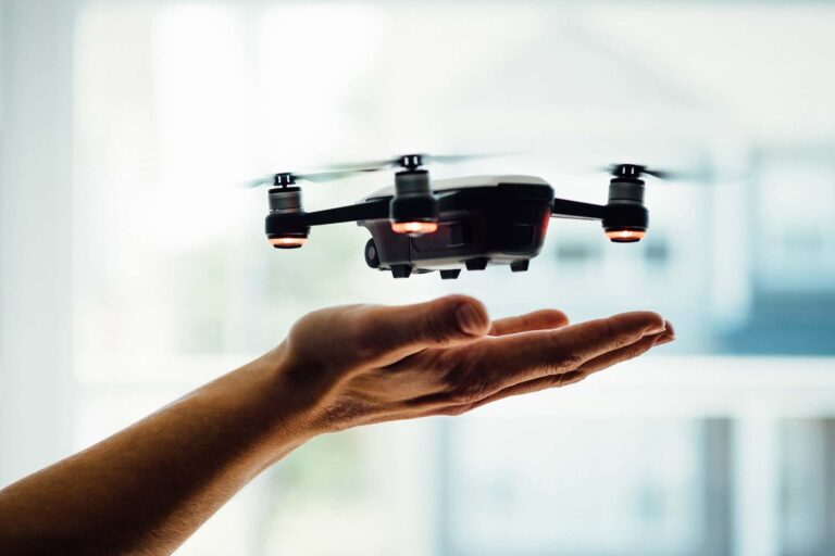 A small drone hovers above an outstretched hand indoors. The background is softly blurred, emphasizing the drone's details and the person's hand beneath it.