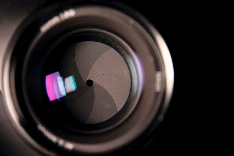 Close-up of a camera lens showing the aperture blades inside. The blades form a circular pattern, and light reflections create colorful hues on the lens surface. The background is black, emphasizing the lens details.