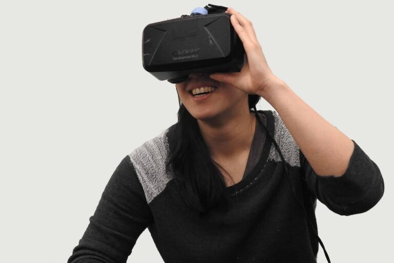 A person wearing a black and gray sweater is smiling while using a virtual reality headset. The background is plain and light-colored.