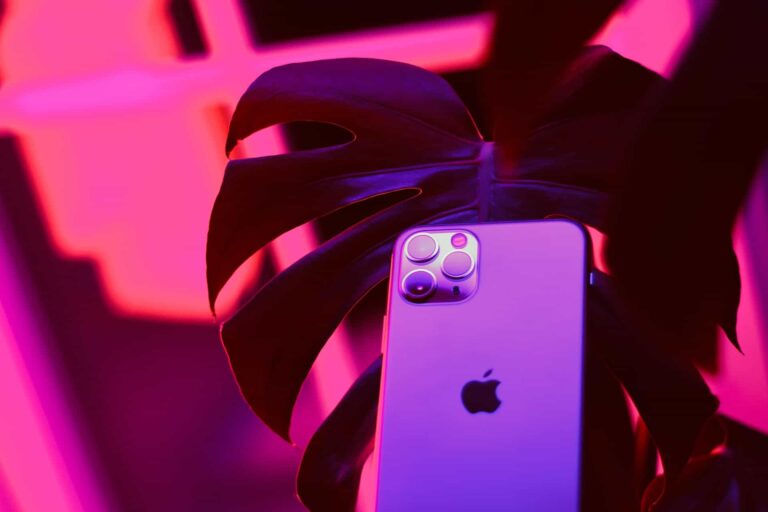 A smartphone with a recognizable apple logo and triple camera setup is partially obscured by a large leaf. The scene is lit with vibrant pink and purple lighting, creating bold shadows and a modern, futuristic feel.