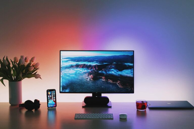 A minimalist desk setup featuring a widescreen monitor displaying a scenic aerial view of clouds. Beside it are a smartphone, headphones, a laptop, a tea cup, a wireless keyboard and mouse, and a vase of tulips, all under colorful ambient lighting.