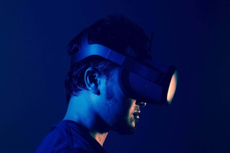 A person wearing a VR headset is shown in profile view against a dark blue background. The headset emits a warm light that illuminates the side of the person's face. The overall atmosphere is futuristic and immersive.