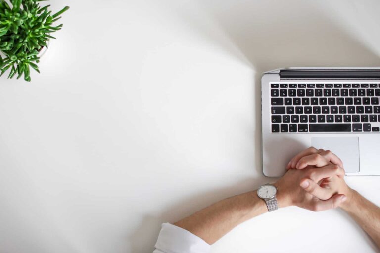 A person with clasped hands rests their forearms on a white desk next to an open laptop. A small green plant in a pot is visible in the top left corner. The scene suggests a minimalist workspace.