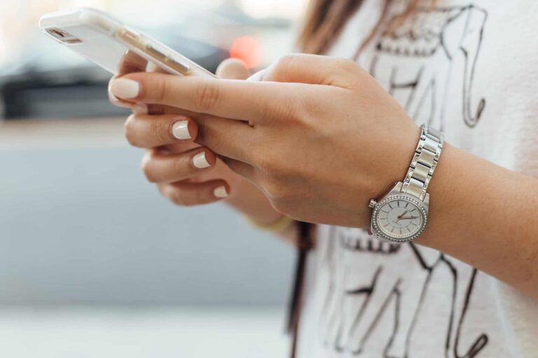 A person wearing a silver wristwatch is holding a smartphone. Their nails are painted white, and they are dressed in a shirt with an illustrated design. The background is blurred.