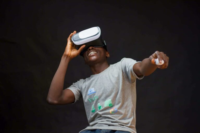 A person wearing a light gray T-shirt is using a virtual reality headset and smiling, with their right arm extended forward. The background is dark, creating contrast with the subject.