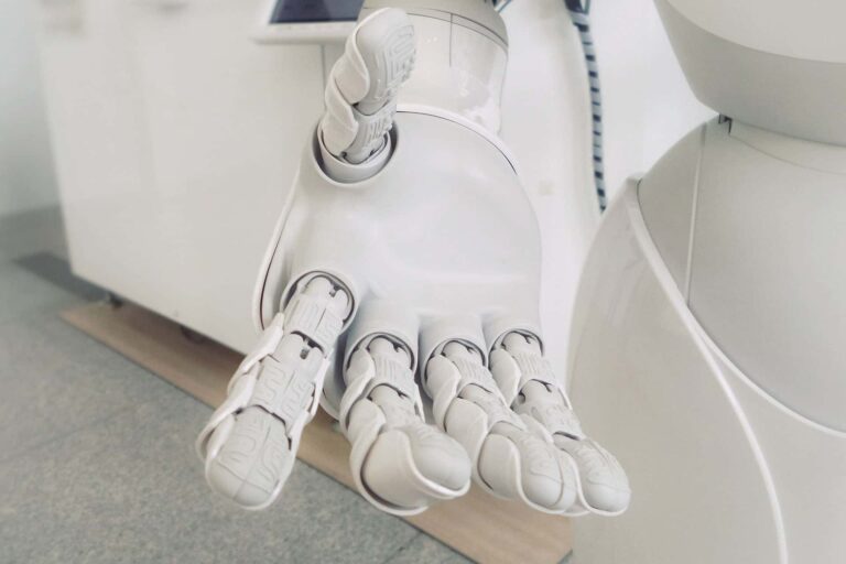 Close-up of a white robotic hand reaching forward, displaying its mechanical fingers and joints. The background is a blurred indoor setting, highlighting the sleek and advanced design of the robot's hand.