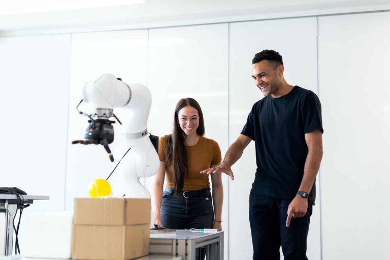 Two people stand in a bright room interacting with a robotic arm on a table. The arm appears to be holding or manipulating an object. Both individuals are smiling and engaged with the technology.