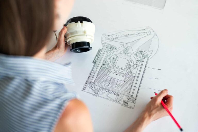 A person holding a white mechanical component and examining detailed technical drawings on a white table. They are wearing a striped sleeveless shirt and holding a red pen, possibly indicating areas on the drawing.