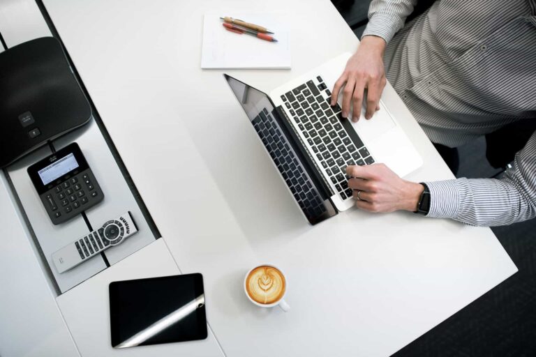 A person typing on a laptop at a white desk with a notebook, pens, and smartphone. A cup of latte and a tablet are nearby, along with an office phone. The person wears a striped shirt and a watch on their left wrist.