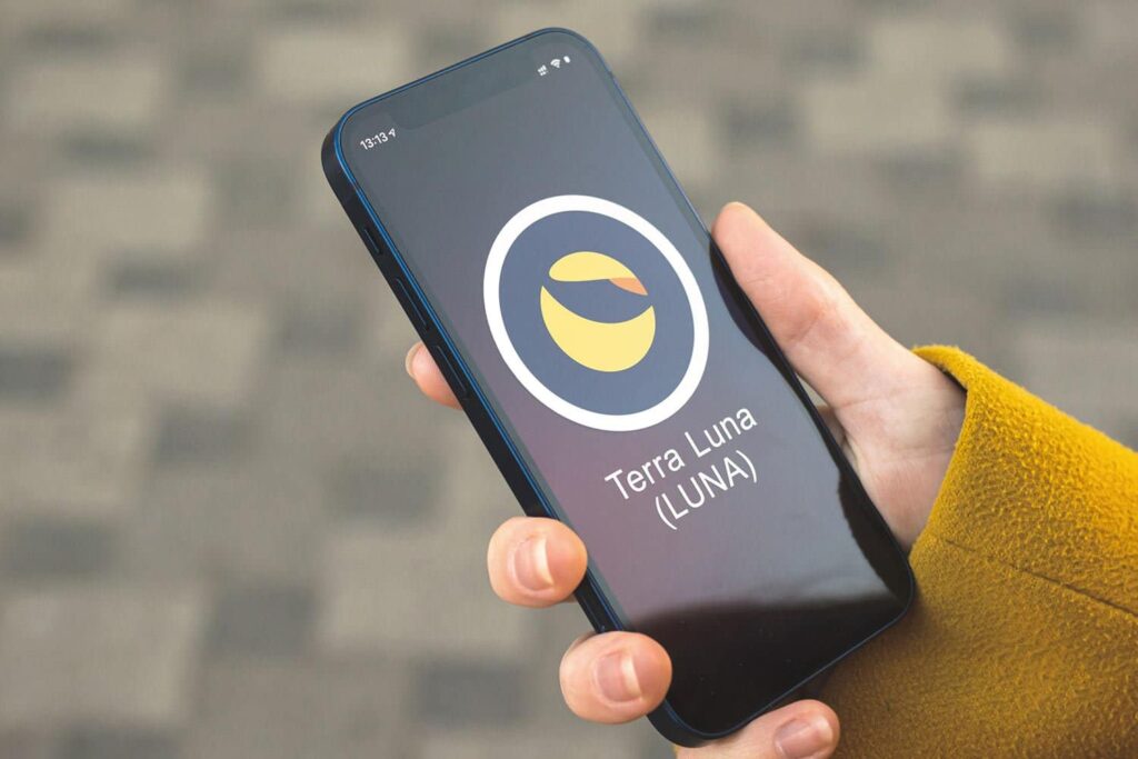 A person holding a smartphone displaying the Terra Luna (LUNA) logo on the screen. The phone's background is a patterned, blurred ground surface, and the person is wearing a yellow coat.