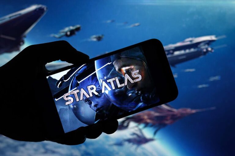 A silhouetted hand holds a smartphone displaying the "Star Atlas" logo with futuristic helmeted figures. In the background, several spacecrafts soar through a starry, cosmic sky.