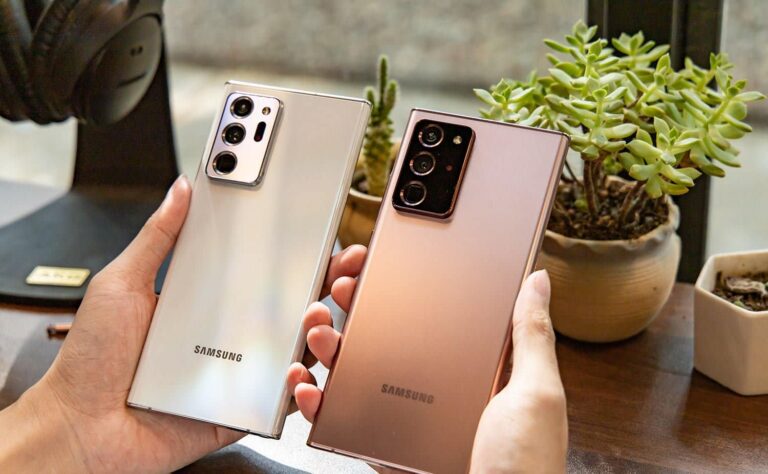 Two hands are holding Samsung smartphones, one silver and one bronze, in front of small potted succulents on a wooden table.