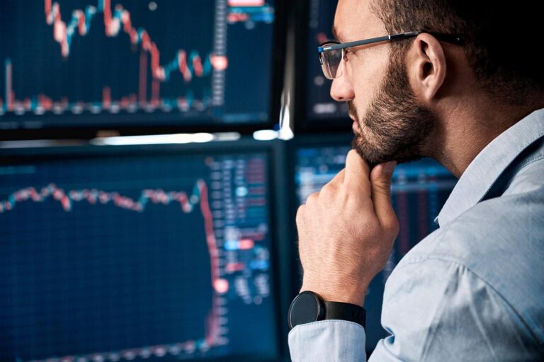 A man in a light blue shirt and glasses is intently analyzing several computer screens displaying stock market graphs and data. He rests his chin on his hand, focusing on the downward market trends shown on the monitors.