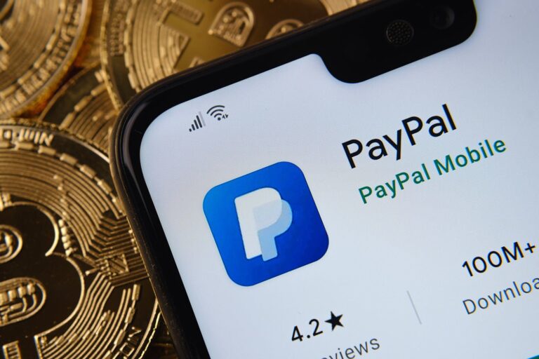 A smartphone screen displays the PayPal app page on a digital store, showing a 4.2-star rating and over 100 million downloads. In the background, there are several Bitcoin coins, hinting at cryptocurrency themes.