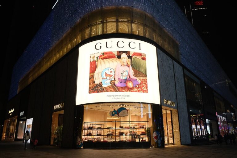 A large Gucci store with an illuminated Doraemon-themed window display. The storefront features a playful scene with Doraemon characters and luxury fashion items. The exterior is sleek with glass and dark panels, and pedestrians are nearby.