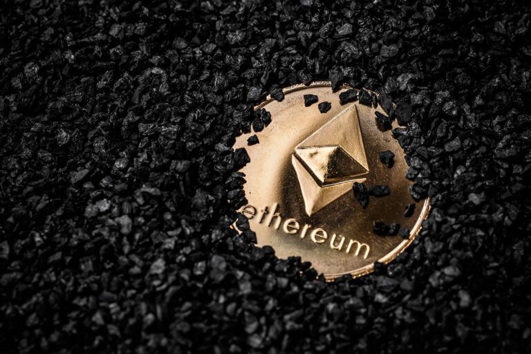 A gold-colored Ethereum coin is partially buried in black gravel. The coin features the Ethereum logo, a geometric shape resembling a diamond, and the word "Ethereum" engraved below it.