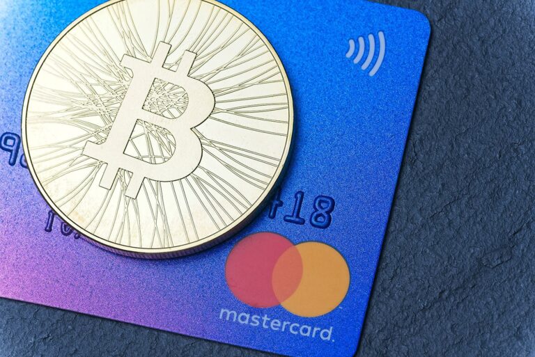 A gold Bitcoin coin rests on a blue gradient Mastercard credit card placed on a dark surface. The Mastercard logo is visible, and the card appears to be equipped with contactless payment technology.