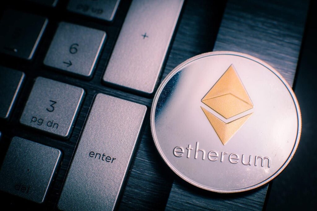 A silver Ethereum coin with a gold symbol is placed on a laptop keyboard, partially covering the "Enter" key. The coin's surface is reflective, capturing light. The keyboard keys are dark with white labels.