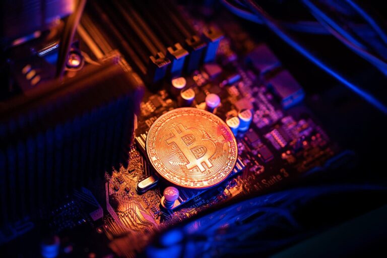 A glowing Bitcoin coin rests on a computer motherboard under dramatic blue and orange lighting, highlighting the intricate circuitry and electronic components surrounding it.